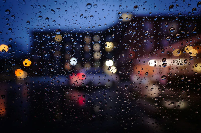 雨のつぶがついた窓ガラスの向こうに、街の光がにじんで見える写真
