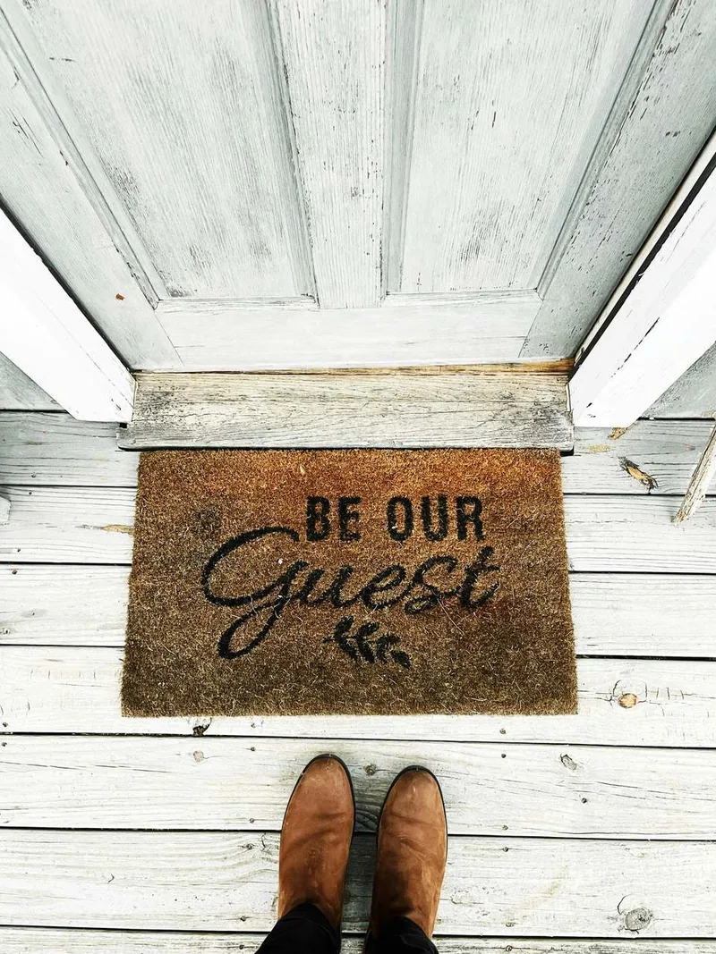 木の戸口に置かれた、「BE OUR GUEST」と書かれた玄関マットの写真